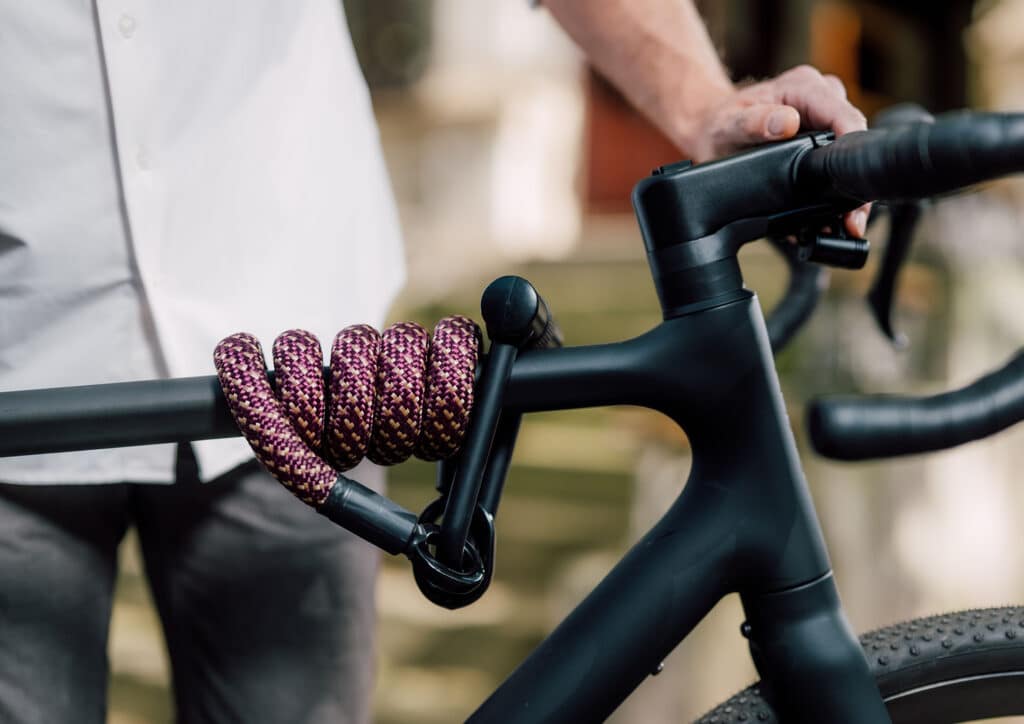 tex–lock eyelet in chateau red mit X-lock flexibel um das Oberrohr vom schwarzen Fahrrad gewickelt, männliches Model im Hintergrund