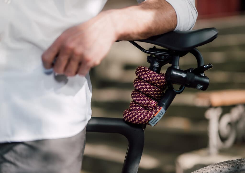 tex–lock eyelet in chateau red mit U-lock flexibel um Sattelstange vom schwarzen Fahrrad gewickelt, männliches Model lehnt dagegen